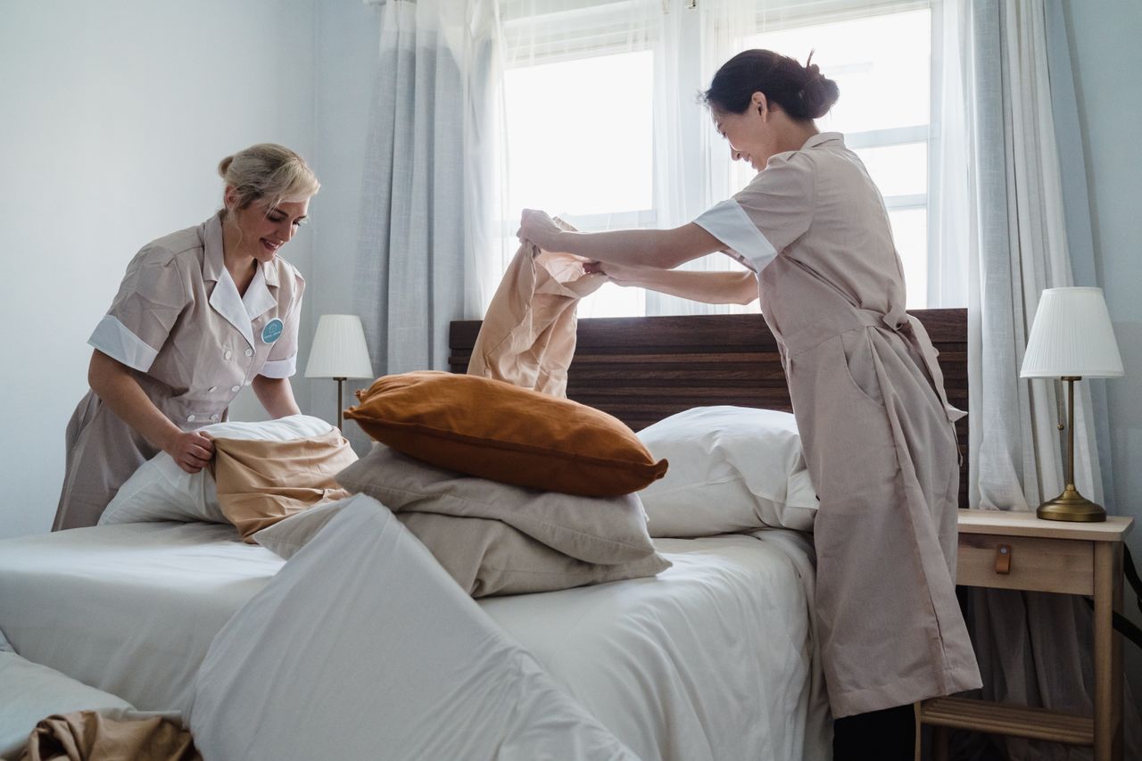 Two housekeepers in uniform making up a hotel bed with crisp linen and pillows.